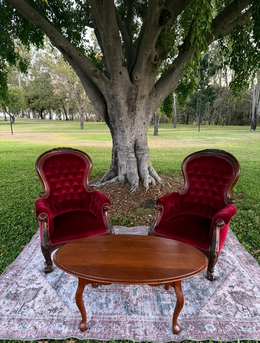 Pair of ruby red velvet high-backed Victorian parlour armchairs with an ornate timber coffee table and vintage rug on a lawn under a large tree, styled as a wedding ceremony signing chair package by Rockhampton Vintage Hire.
